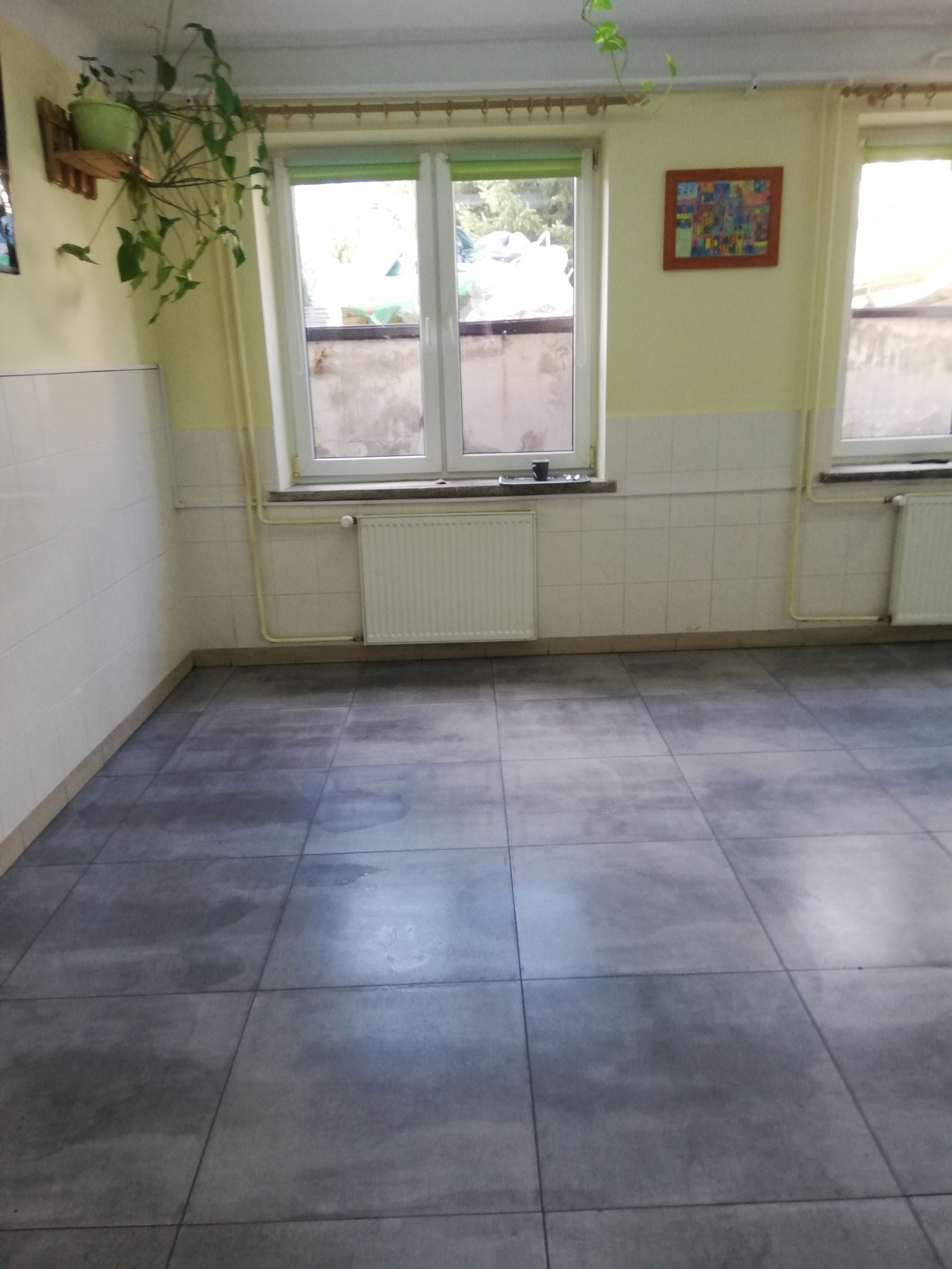 Wnętrze pokoju z szarymi płytkami podłogowymi, białymi płytkami na ścianach do połowy wysokości, dwoma oknami z zielonymi roletami i grzejnikami pod parapetami, oraz rośliną doniczkową na ścianie...