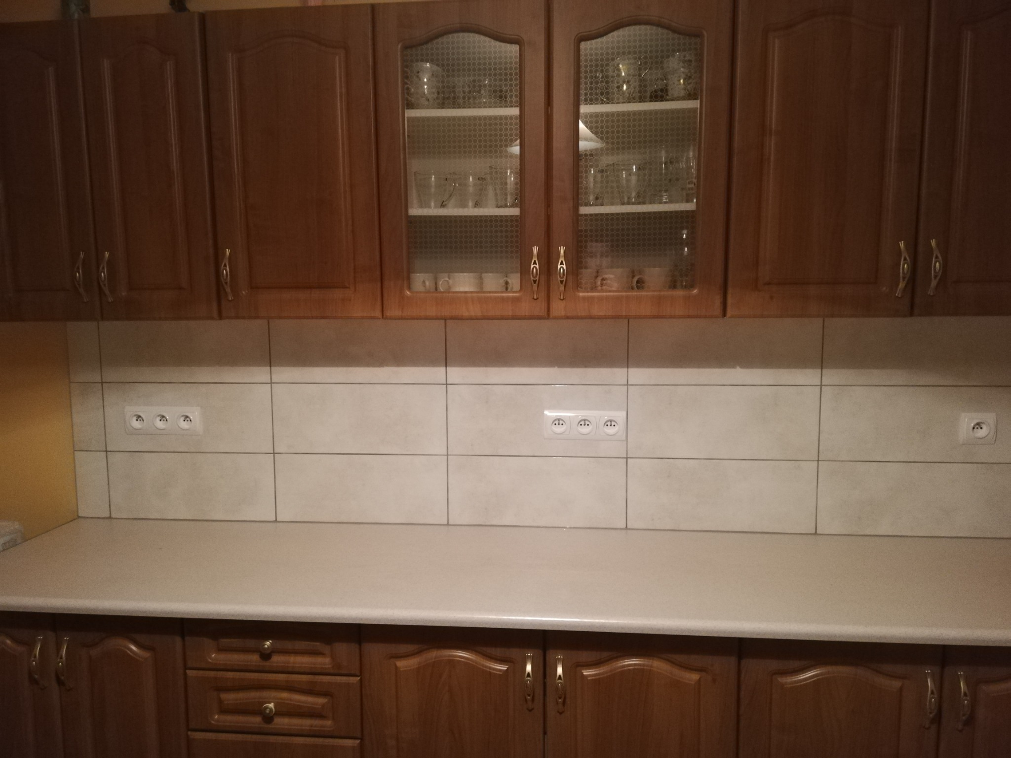 Klasyczna kuchnia z drewnianymi szafkami w odcieniach brązu, jasnym blatem i beżowymi płytkami ściennymi z widocznymi gniazdkami elektrycznymi.