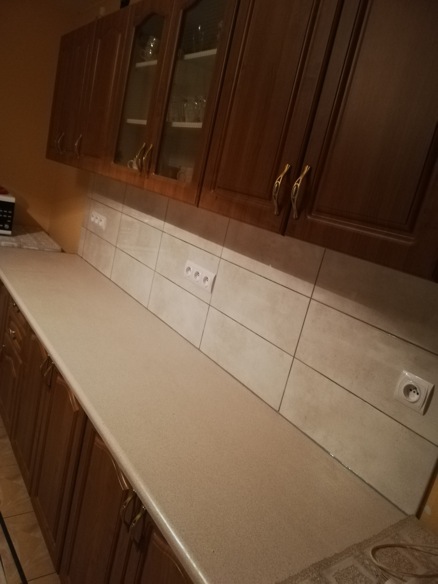 Wykończona kuchnia z brązowymi szafkami, beżowym blatem i jasnymi, kwadratowymi płytkami na ścianie, widoczne gniazdka elektryczne.