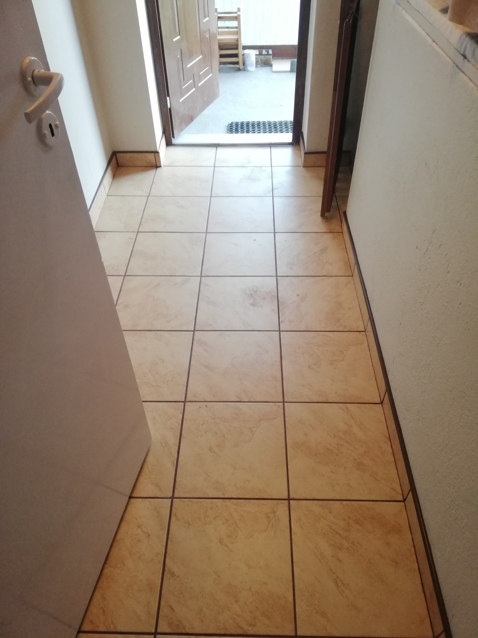 Beżowe, kwadratowe płytki podłogowe, ułożone w korytarzu, widoczne drzwi wejściowe i fragment ściany.