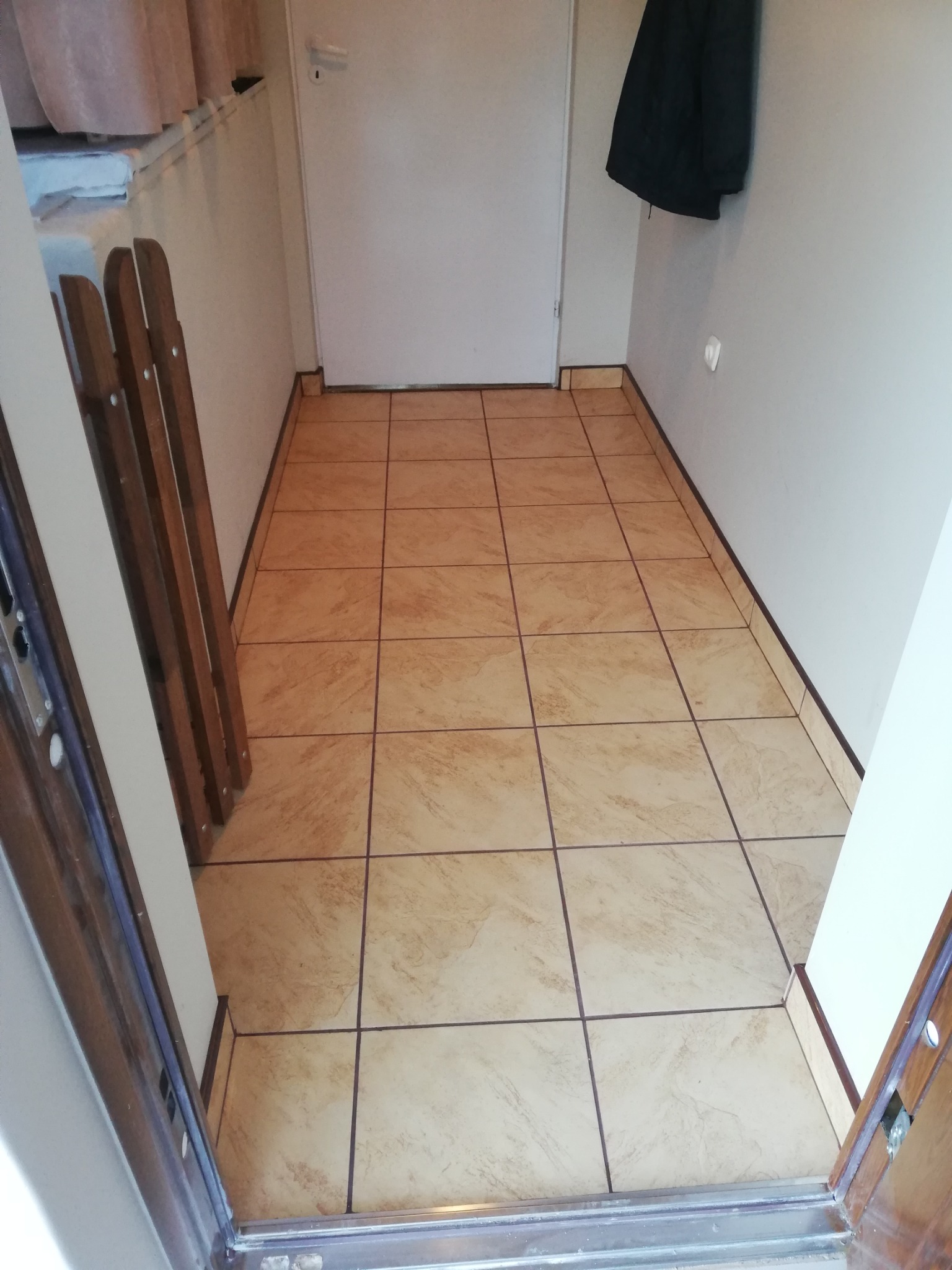 Beżowe, kwadratowe płytki podłogowe z ciemniejszymi fugami w korytarzu, widoczne z perspektywy wejścia, z drewnianą barierką po lewej stronie i kurtką wiszącą na ścianie po prawej.