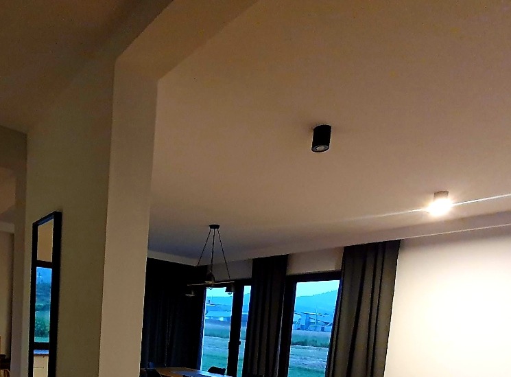 Nowoczesne oświetlenie sufitowe w salonie z dużymi oknami i ciemnymi zasłonami, widok na zewnątrz w pochmurny dzień.