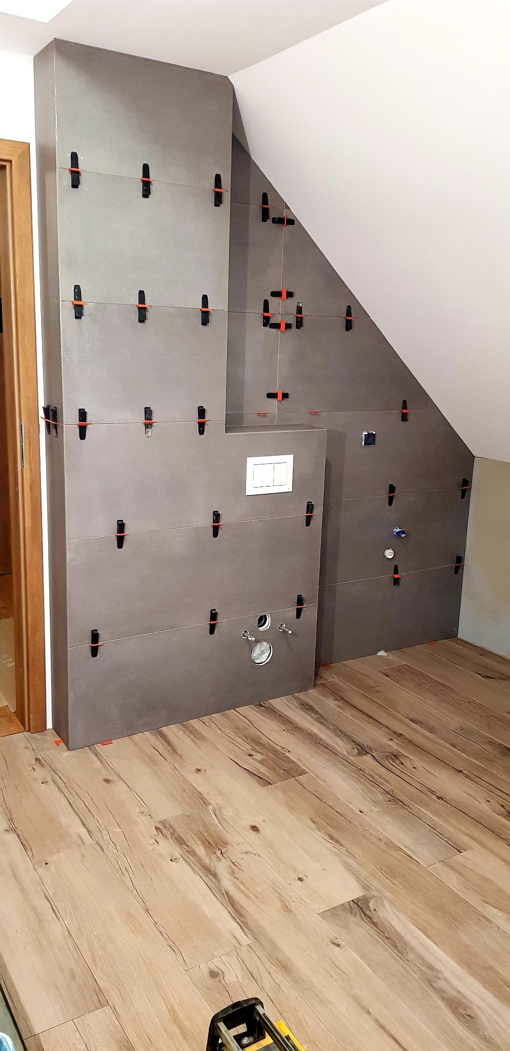 Szare płytki ścienne w trakcie montażu w łazience ze skosem, widoczne systemy poziomowania płytek, przygotowane otwory na instalacje, drewnopodobna podłoga