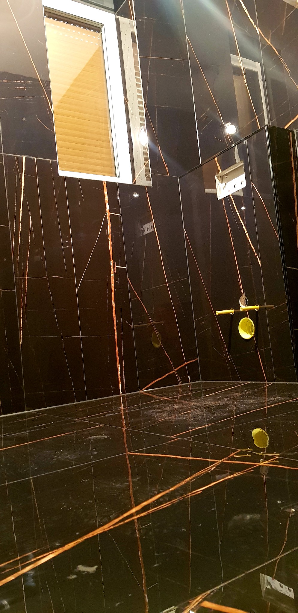 Ciemne płytki imitujące marmur z wyraźnymi złotymi żyłami na ścianie i podłodze łazienki, widoczny fragment okna z żaluzjami i przygotowane miejsce na armaturę.
