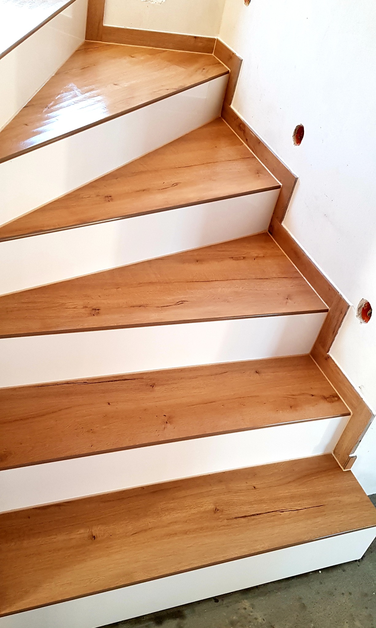 Wykończone schody z drewnopodobnymi płytkami i białymi podstopnicami, widoczne otwory na gniazdka elektryczne w ścianie.