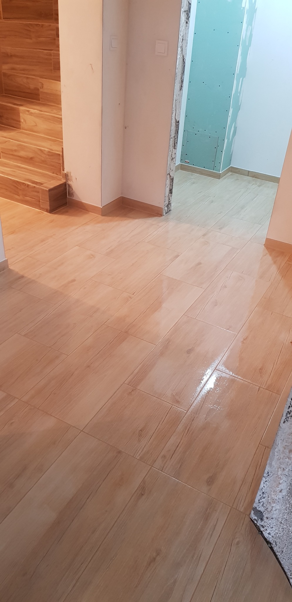 Ułożona podłoga z płytek imitujących drewno w nowym budynku, widoczne schody również wykończone płytkami, ściany przygotowane do malowania.