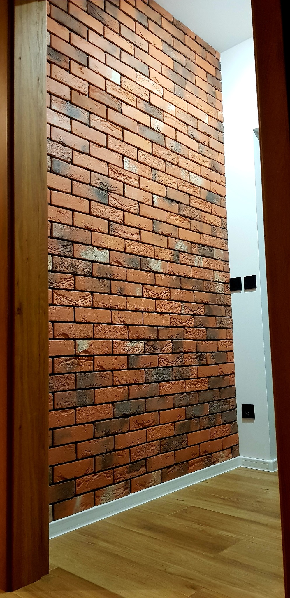 Ściana wyłożona cegłą klinkierową w odcieniach czerwieni i brązu, widoczna z perspektywy wejścia do pomieszczenia, z drewnianą framugą po lewej stronie i białymi ścianami z czarnymi gniazdkami po...