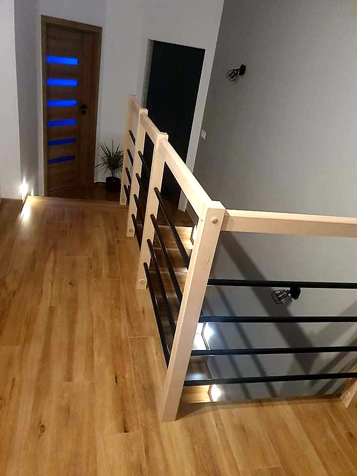 Drewniana podłoga z widoczną klatką schodową z jasnego drewna i czarnymi metalowymi elementami. W tle widoczne drzwi z niebieskimi szybami i oświetlenie podłogowe.