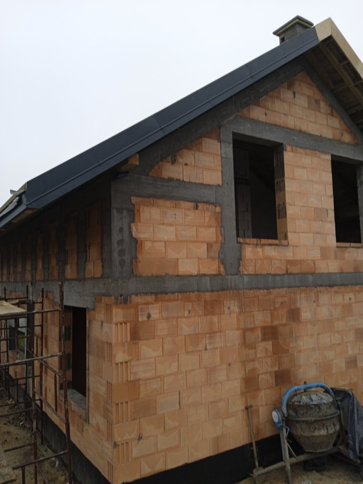 Surowy stan domu z cegły ceramicznej z ciemnym dachem, widoczne otwory okienne, zacierki betonowe i stojąca betoniarka.