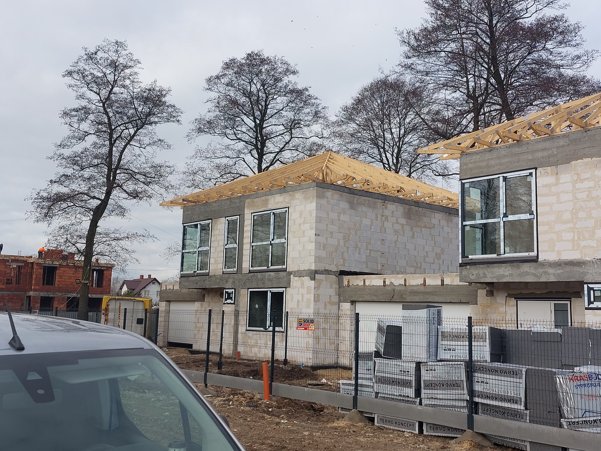 Dwa domy w stanie surowym otwartym, z widoczną konstrukcją drewnianą dachu, okna zamontowane, ściany z betonu komórkowego, w tle budynek w trakcie murowania ścian z cegły, teren budowy...