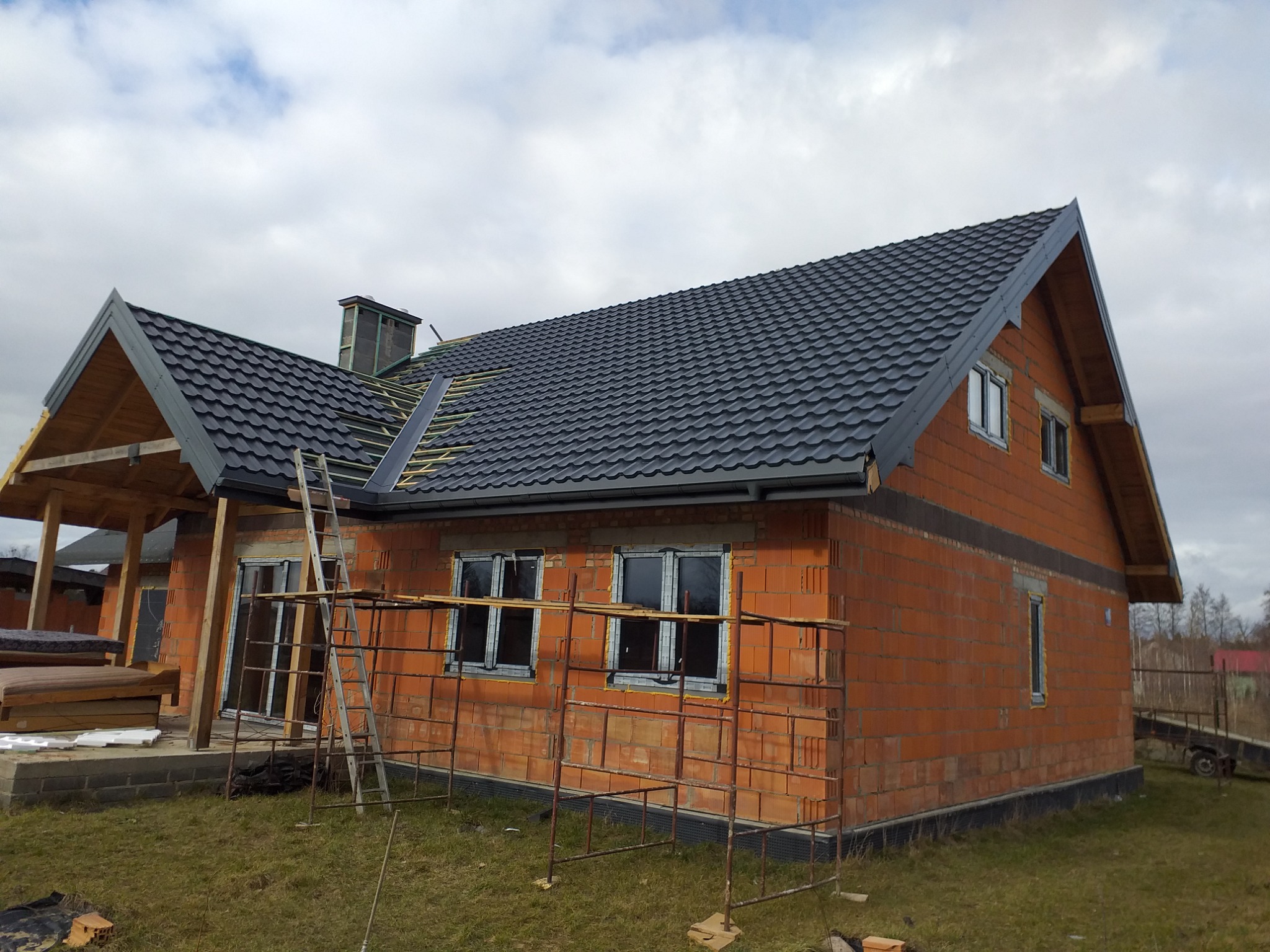 Dom w trakcie budowy z czerwonej cegły, częściowo pokryty ciemnoszarą dachówką, widoczne rusztowania i drabina, fragmenty więźby dachowej bez pokrycia.