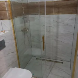 Nowoczesna kabina prysznicowa z złotymi ramami i odpływem liniowym, marmurowe płytki na ścianach i podłodze, wisząca toaleta w rogu.