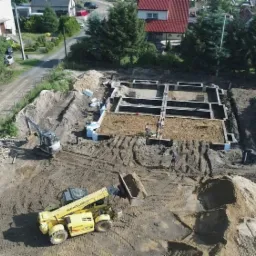 Widok z góry na plac budowy z wykopanymi fundamentami pod dom, gdzie pracują robotnicy i maszyny budowlane: koparka i ładowarka teleskopowa.