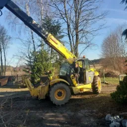 Żółta ładowarka teleskopowa New Holland z operatorem w kabinie, na błotnistym terenie budowy, z podniesionym ramieniem, widoczne drzewa w tle i niebieskie niebo.