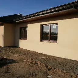 Nowo otynkowany budynek jednorodzinny z brązowymi oknami i ciemną dachówką, widoczne ślady prac ziemnych wokół budynku.