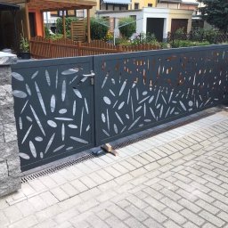 TGA FIJOŁEK Sp. z o.o. - Nowoczesna, przesuwna brama w kolorze grafitowym z ażurowym wzorem liści, zamontowana na posesji z kostki brukowej, widoczne słupki z szarego kamienia i drewniane ogrodzenie w tle.