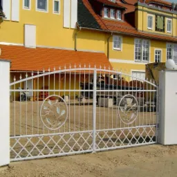 Biała, metalowa brama wjazdowa z motywem liści dębu w okręgach, zamontowana na białych, murowanych słupkach z kulami, na tle żółtego budynku z pomarańczową dachówką.