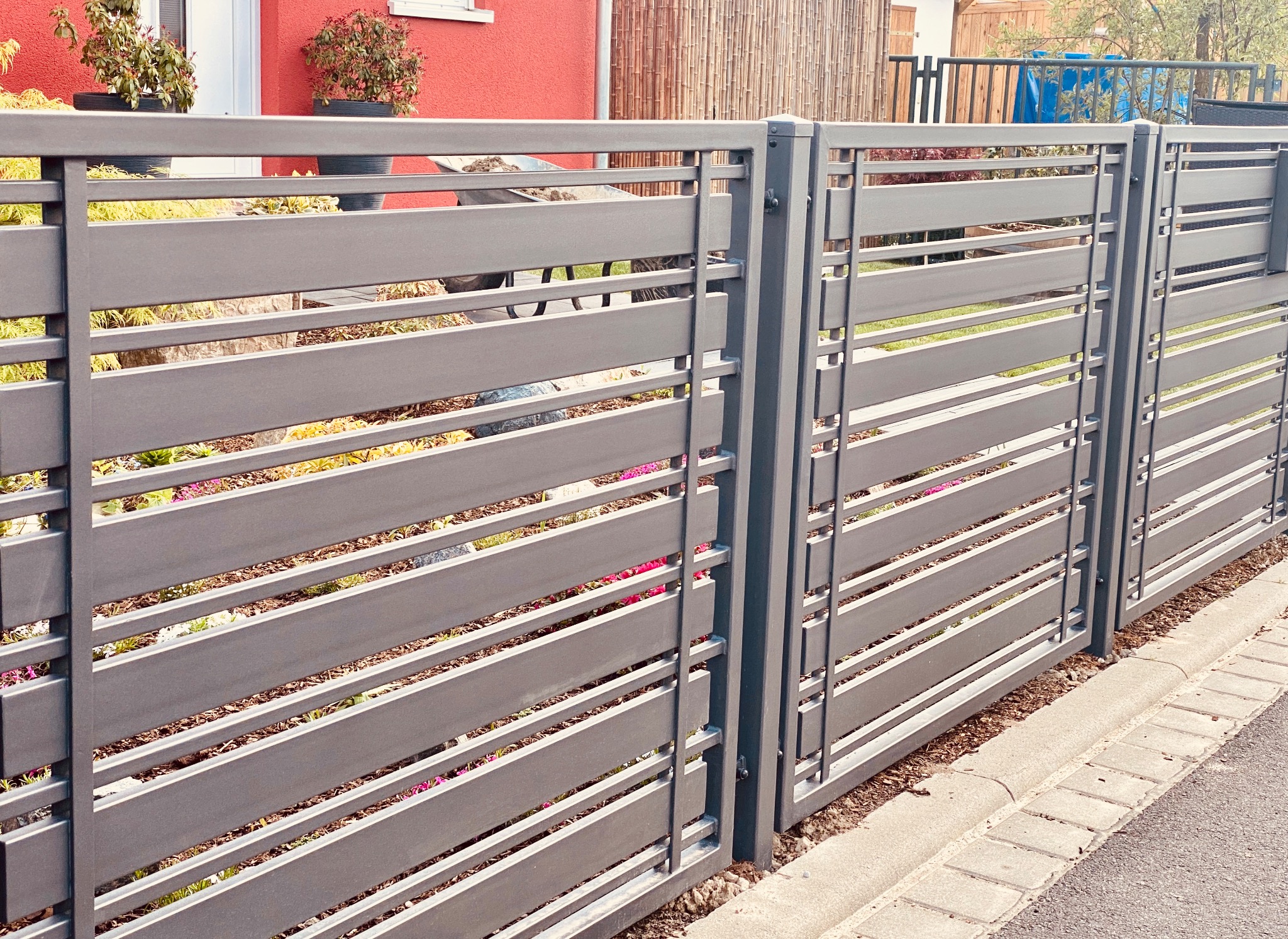 Nowoczesne, poziome ogrodzenie panelowe w kolorze szarym, z widocznymi elementami montażowymi i roślinnością w tle, ustawione wzdłuż betonowego obrzeża.