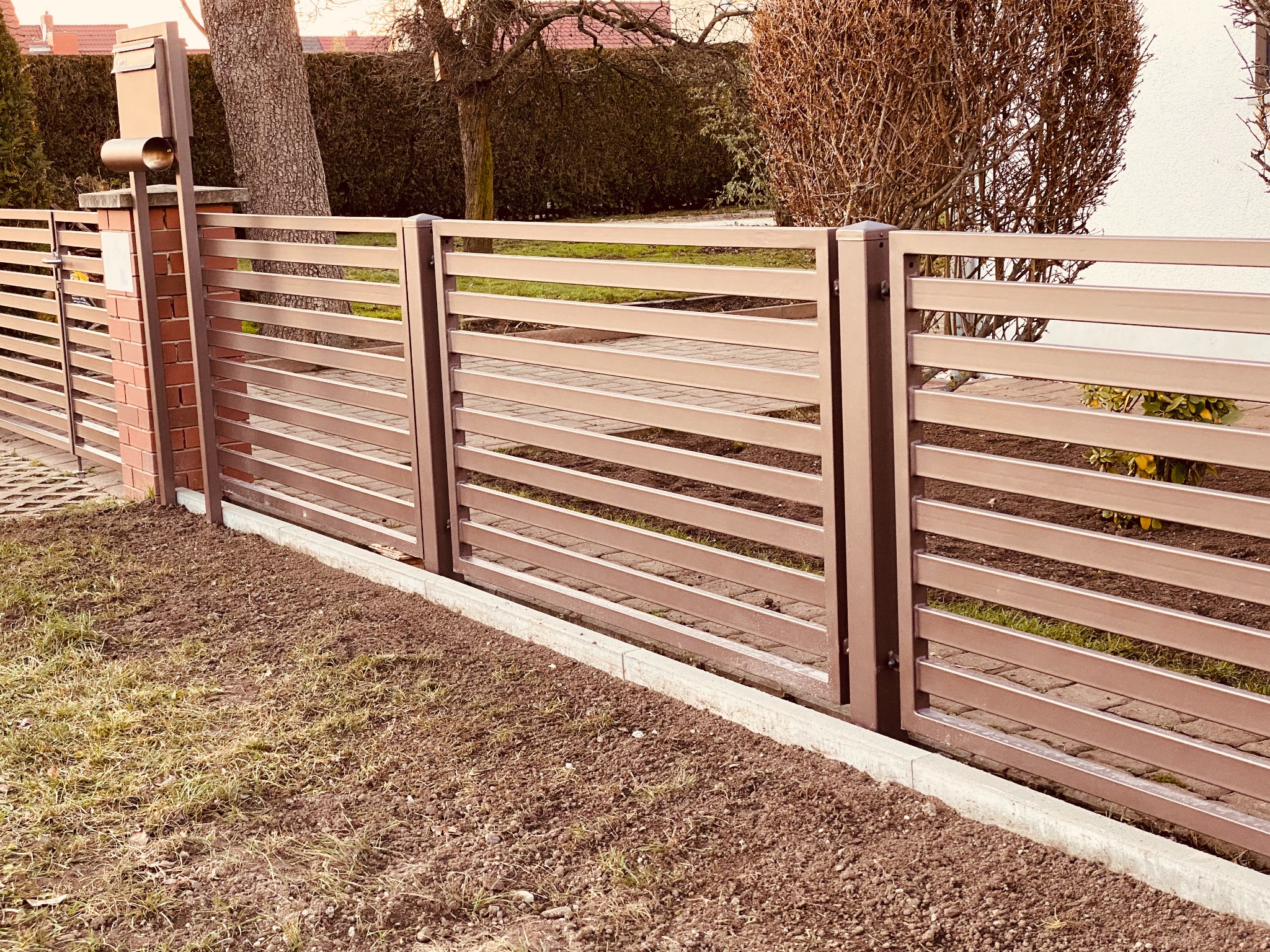 Nowoczesne ogrodzenie panelowe w kolorze brązowym z poziomymi przęsłami, zamontowane na ceglanym słupku z metalową skrzynką na listy, widok z niskiej perspektywy na trawnik i świeżo przekopaną ziemię.