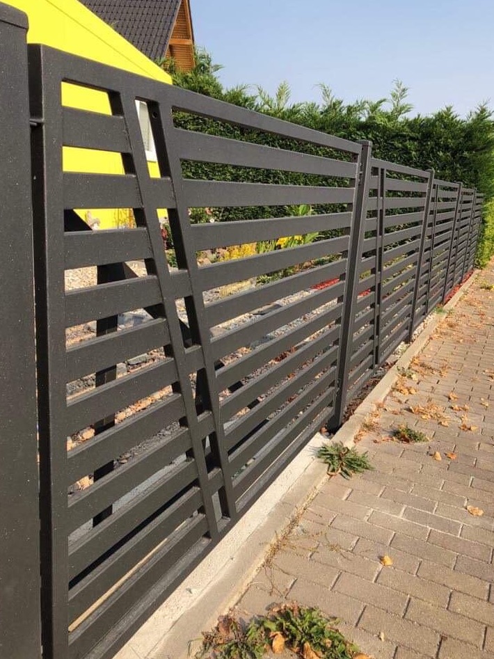 Nowoczesne, poziome przęsła ogrodzeniowe w kolorze antracytowym, z ukośnymi wzmocnieniami, oddzielające posesję od chodnika z kostki brukowej, na tle zielonego żywopłotu i żółtego budynku.