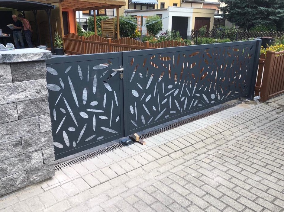 Nowoczesna, przesuwna brama w kolorze grafitowym z ażurowym wzorem liści, zamontowana na posesji z kostki brukowej, widoczne słupki z szarego kamienia i drewniane ogrodzenie w tle.