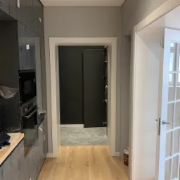 Wnętrze mieszkania w trakcie remontu: zabudowa kuchenna z folią ochronną, podłoga z jasnego drewna, widok na pomieszczenie z szarą podłogą w kształcie plastra miodu i czarną ścianą, białe drzwi...