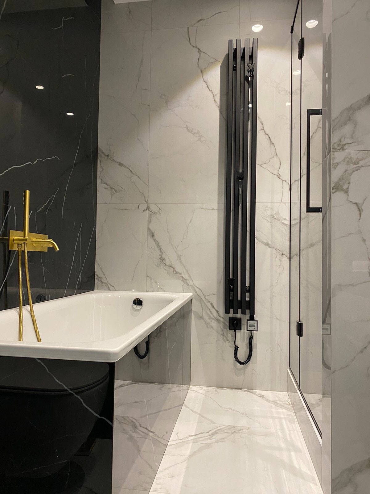 Elegancka łazienka z czarno-białymi marmurowymi płytkami, wolnostojącą wanną z czarną obudową i złotą armaturą, nowoczesnym czarnym grzejnikiem drabinkowym i kabiną prysznicową ze szklanymi drzwiami.