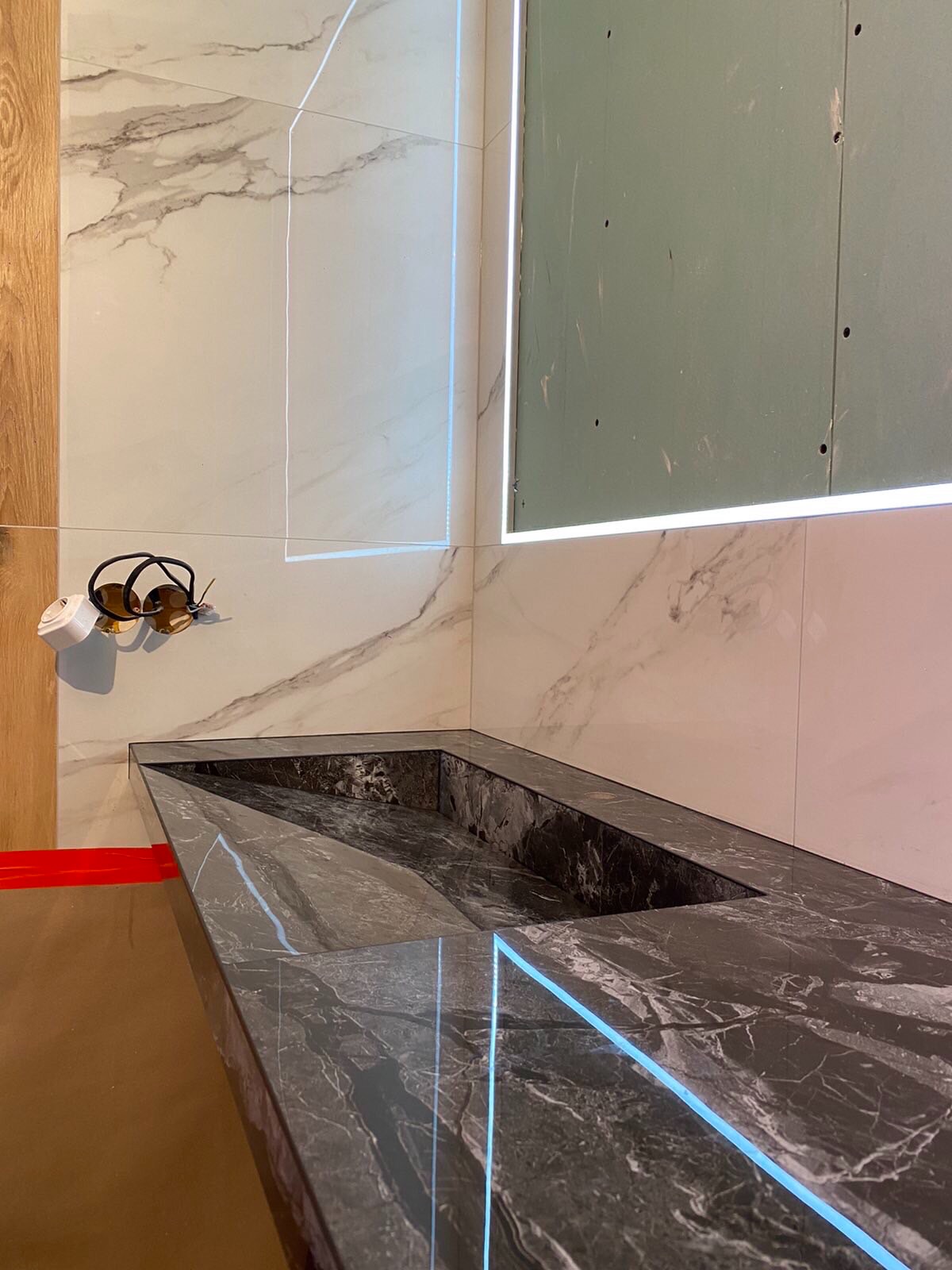 Marmurowa umywalka wpuszczona w blat, podświetlona listwą LED, w łazience z białymi marmurowymi płytkami, widoczne przygotowania do montażu gniazdka elektrycznego.