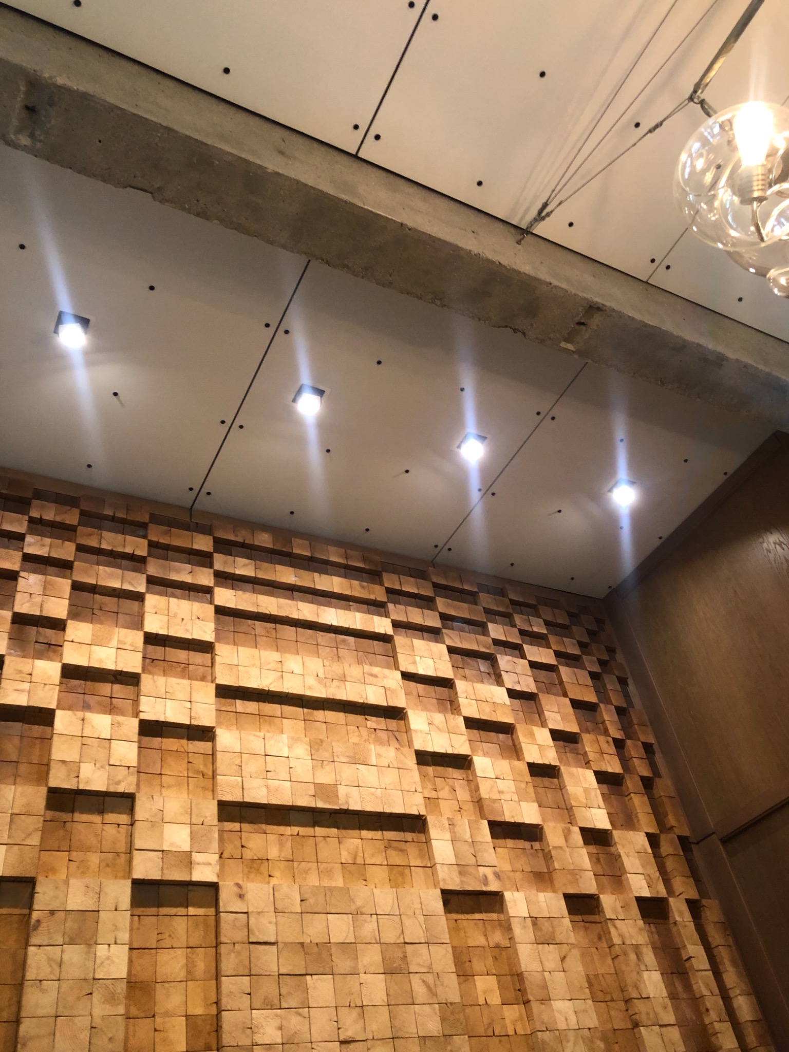 Ściana z drewnianych kostek tworzących trójwymiarowy wzór, oświetlona kwadratowymi lampami sufitowymi i ozdobną lampą wiszącą.