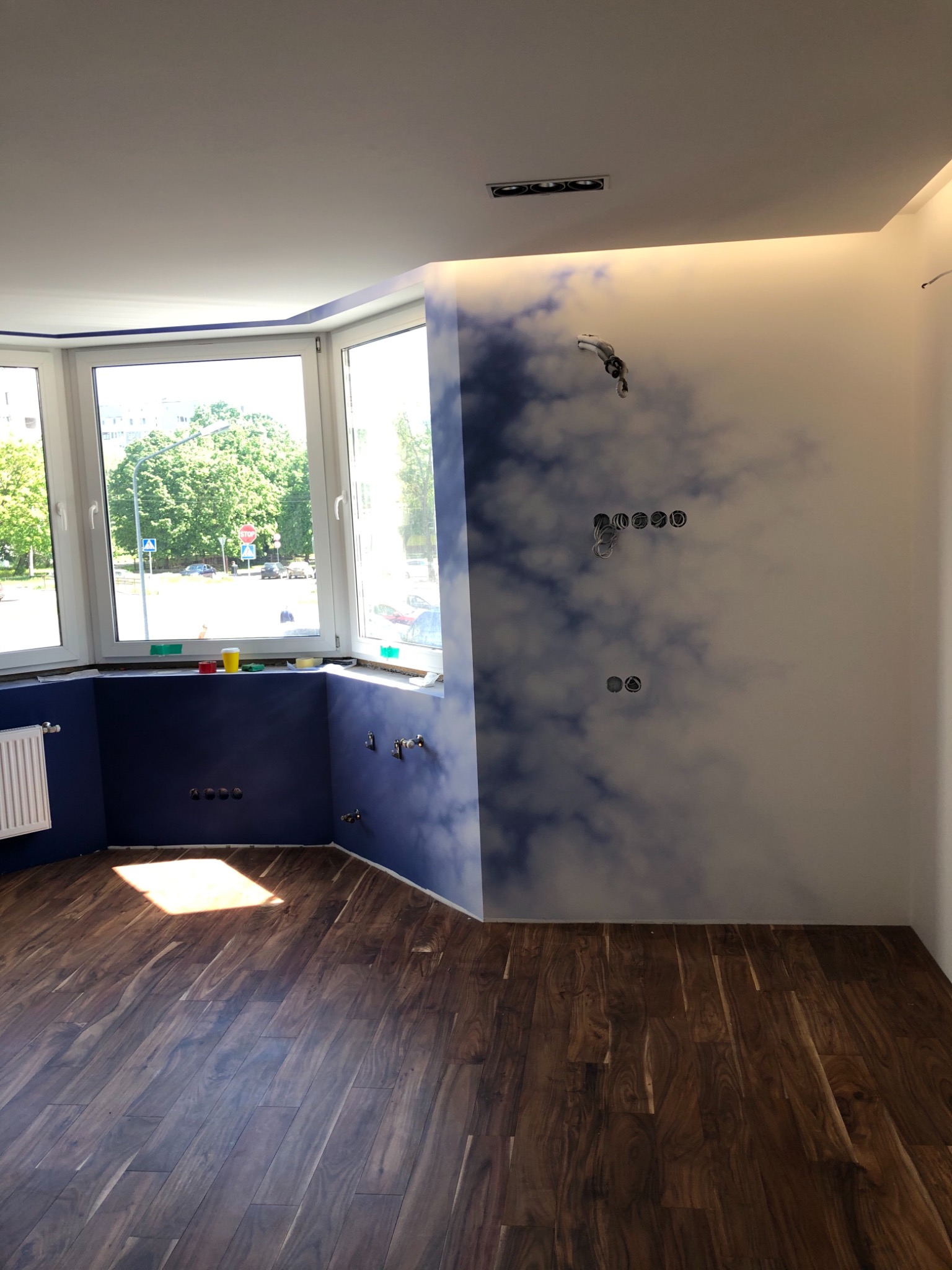 Pokój w trakcie wykańczania: ciemna podłoga, ściany z motywem chmur, okno wykuszowe z widokiem na zieleń, widoczne puszki elektryczne i rury hydrauliczne.