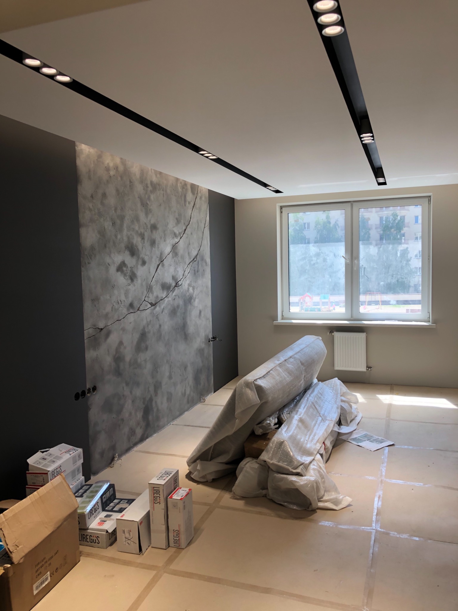 Pokój w trakcie prac wykończeniowych: ściana z efektem betonu z wyraźnym pęknięciem, czarne, liniowe oświetlenie sufitowe, okno z widokiem na plac zabaw, podłoga zabezpieczona folią, stosy...