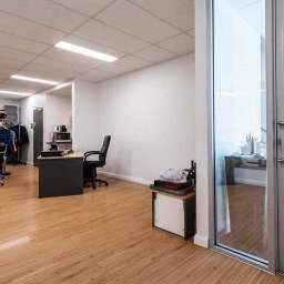 Green & Clean - Jasne biuro z drewnianą podłogą, biurkami i krzesłami. Widoczne przeszklone drzwi i białe ściany. Minimalistyczny design z elementami wyposażenia biurowego.