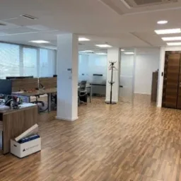 Jasne, przestronne biuro z drewnianą podłogą, biurkami z monitorami i krzesłami, widoczne białe ściany, przeszklone ścianki działowe, w tle wieszak na ubrania oraz brązowe drzwi ewakuacyjne...