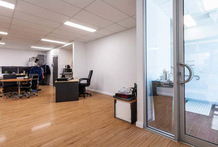 Jasne biuro z drewnianą podłogą, biurkami i krzesłami. Widoczne przeszklone drzwi i białe ściany. Minimalistyczny design z elementami wyposażenia biurowego.