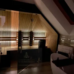 Nowoczesna sauna fińska z przeszklonymi drzwiami w skosie poddasza, z widocznym piecem i oświetleniem LED, obok wanna wolnostojąca z akcesoriami.