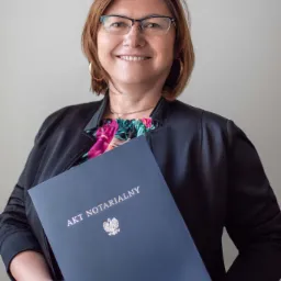 Uśmiechnięta kobieta w okularach trzyma w dłoniach akt notarialny z widocznym godłem Polski i danymi kancelarii.