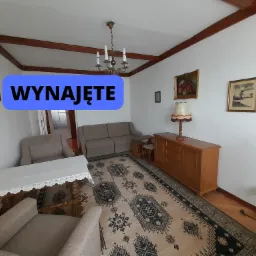 Umeblowane wnętrze pokoju z drewnianymi belkami stropowymi, dywanem w orientalne wzory, brązową komodą, stołem z koronkowym obrusem oraz kanapą i fotelami w odcieniach beżu. Na ścianach obrazy...