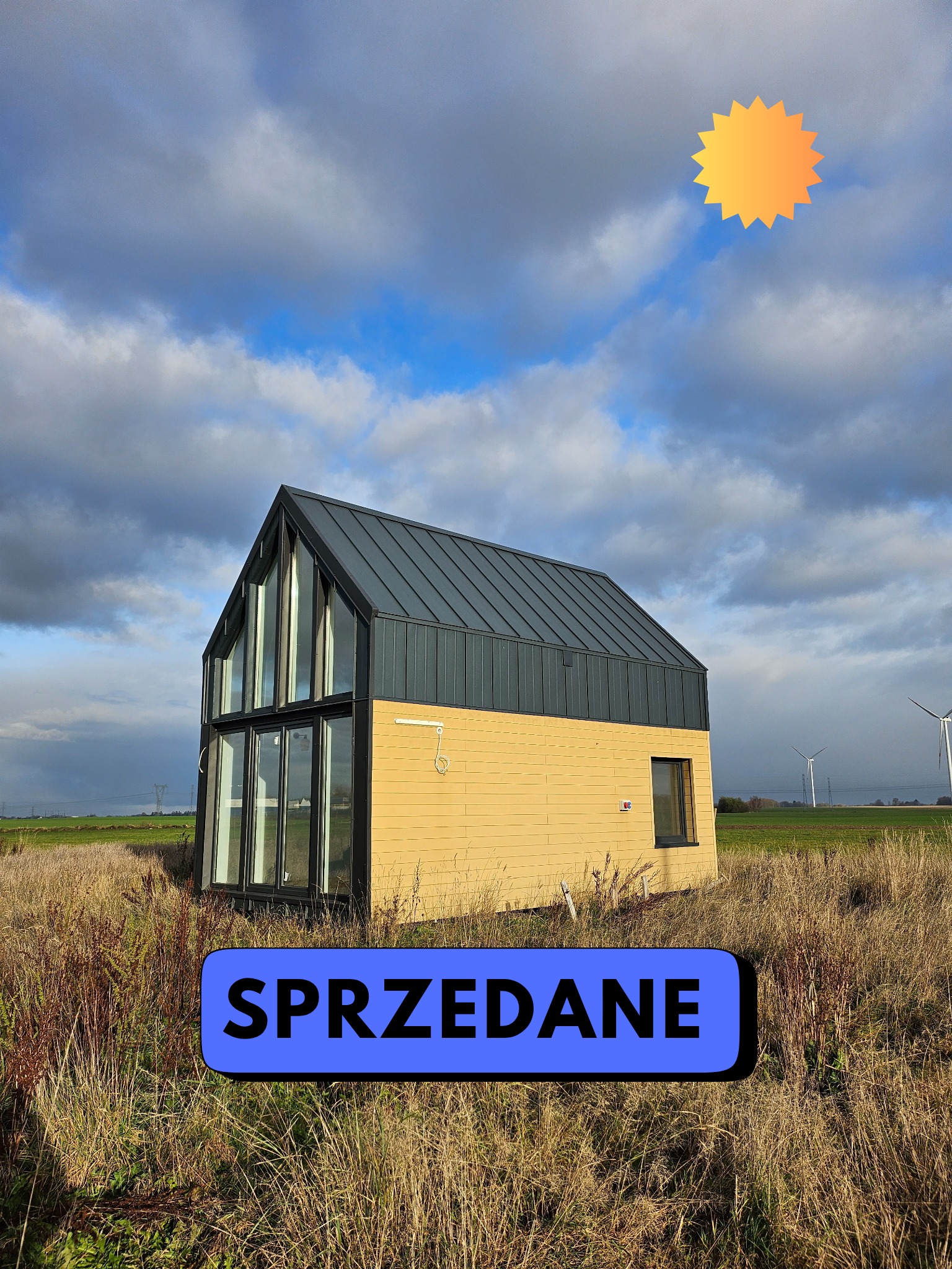 Nowoczesny, żółty dom z ciemnym dachem i dużymi oknami, otoczony suchą trawą. W tle turbiny wiatrowe i pochmurne niebo. Na zdjęciu widnieje napis 'Sprzedane'.