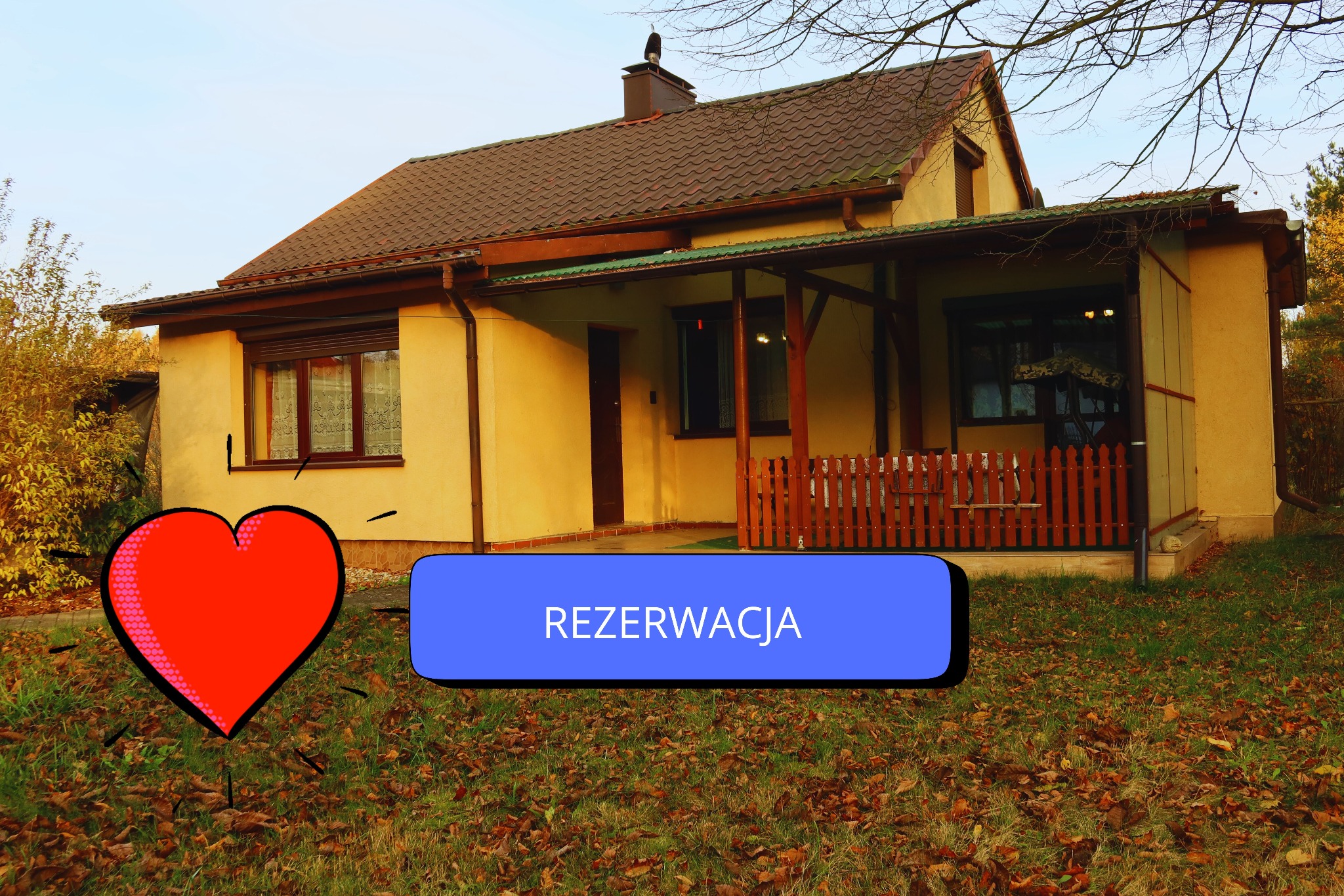 Parterowy dom z brązowym dachem, werandą i napisem 'Rezerwacja' na trawniku. Dodatkowo grafika serca. Ujęcie w jesiennej scenerii, z drzewami w tle.