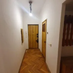 Korytarz z drewnianą podłogą w jodełkę, świeżo odnowione białe ściany, widoczne dwie drewniane drzwi i wisząca lampa z żar&oacute;wką.