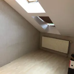 Poddasze po remoncie, ściany pokryte jasną tapetą o delikatnej fakturze, drewniana podłoga, biały grzejnik pod oknem dachowym z częściowo zasłoniętą roletą.