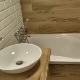 Biała umywalka nablatowa z chromowaną baterią, obok wanna z baterią prysznicową, ściany wyłożone białymi i drewnopodobnymi płytkami.