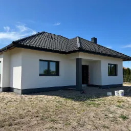 Nowoczesny, parterowy dom z białą elewacją i ciemną dachówką, widok z zewnątrz w słoneczny dzień, teren wokół domu jeszcze niezagospodarowany, widoczne puszki po farbie.