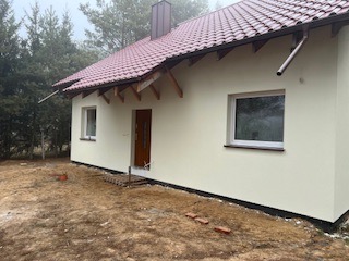 Nowo otynkowany dom jednorodzinny z czerwonym dachem i brązowymi rynnami, widok z boku, teren wokół domu częściowo niezagospodarowany, fragmenty cegieł na ziemi.