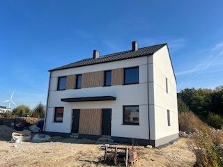 Nowoczesny, dwukondygnacyjny dom bliźniak z jasną elewacją i elementami dekoracyjnymi z drewna na parterze i pierwszym piętrze, czarny dach, teren wokół domu w trakcie prac wykończeniowych...