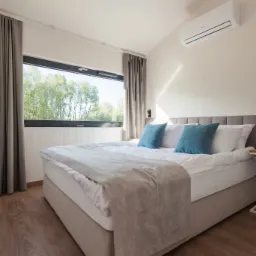 Jasna sypialnia z dużym oknem z widokiem na drzewa, łóżko z szarym zagłówkiem i turkusowymi poduszkami, klimatyzator na ścianie, drewniana podłoga.