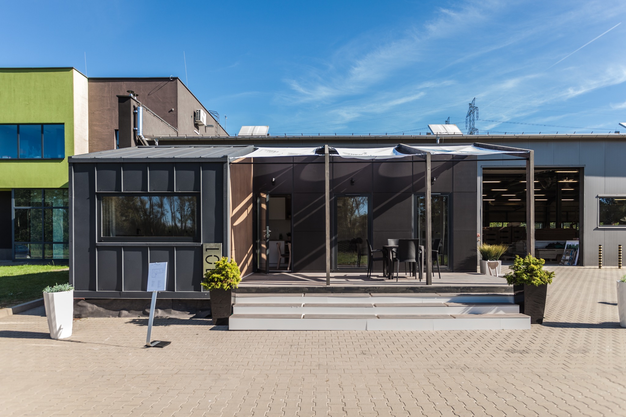 Nowoczesny, parterowy budynek z ciemnoszarą elewacją, białymi schodami wejściowymi i roślinami w donicach, prezentowany na tle błękitnego nieba i innych budynków o zróżnicowanej kolorystyce.