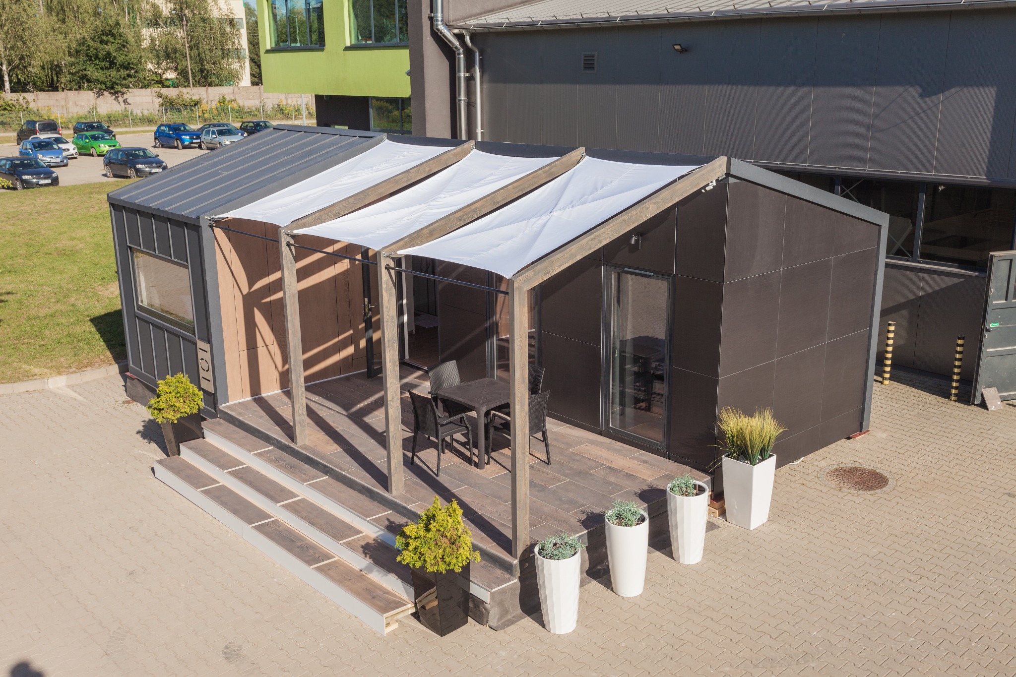 Nowoczesny dom modułowy z zadaszonym tarasem, wyposażonym w stół i krzesła, otoczony roślinami w donicach, na tle budynku przemysłowego i parkingu.