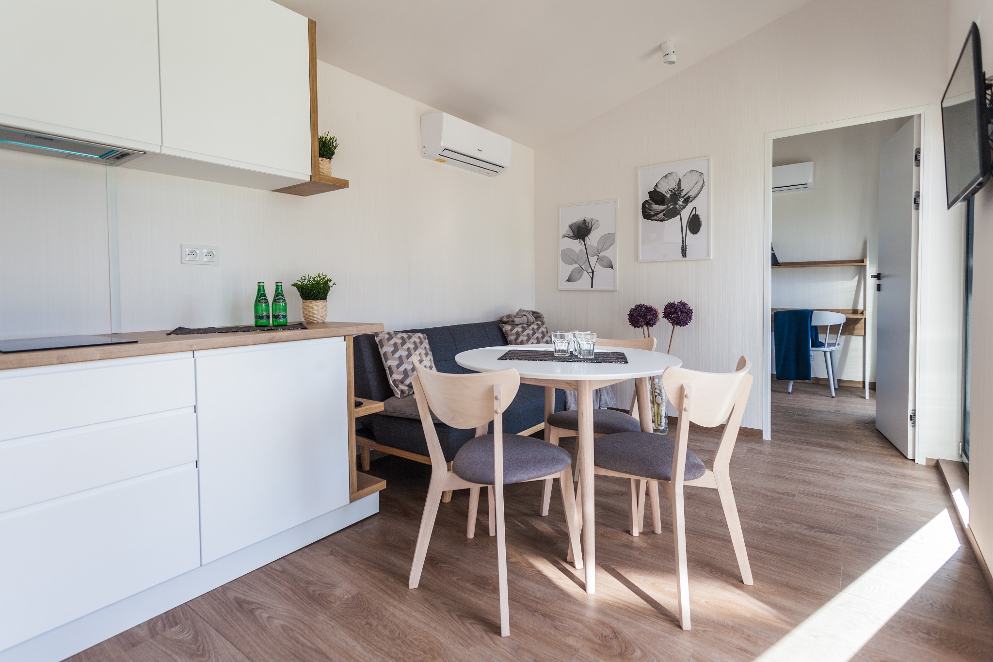 Jasne wnętrze aneksu kuchennego z okrągłym stołem i krzesłami w stylu skandynawskim; widoczne dekoracje, klimatyzacja i wejście do kolejnego pomieszczenia.