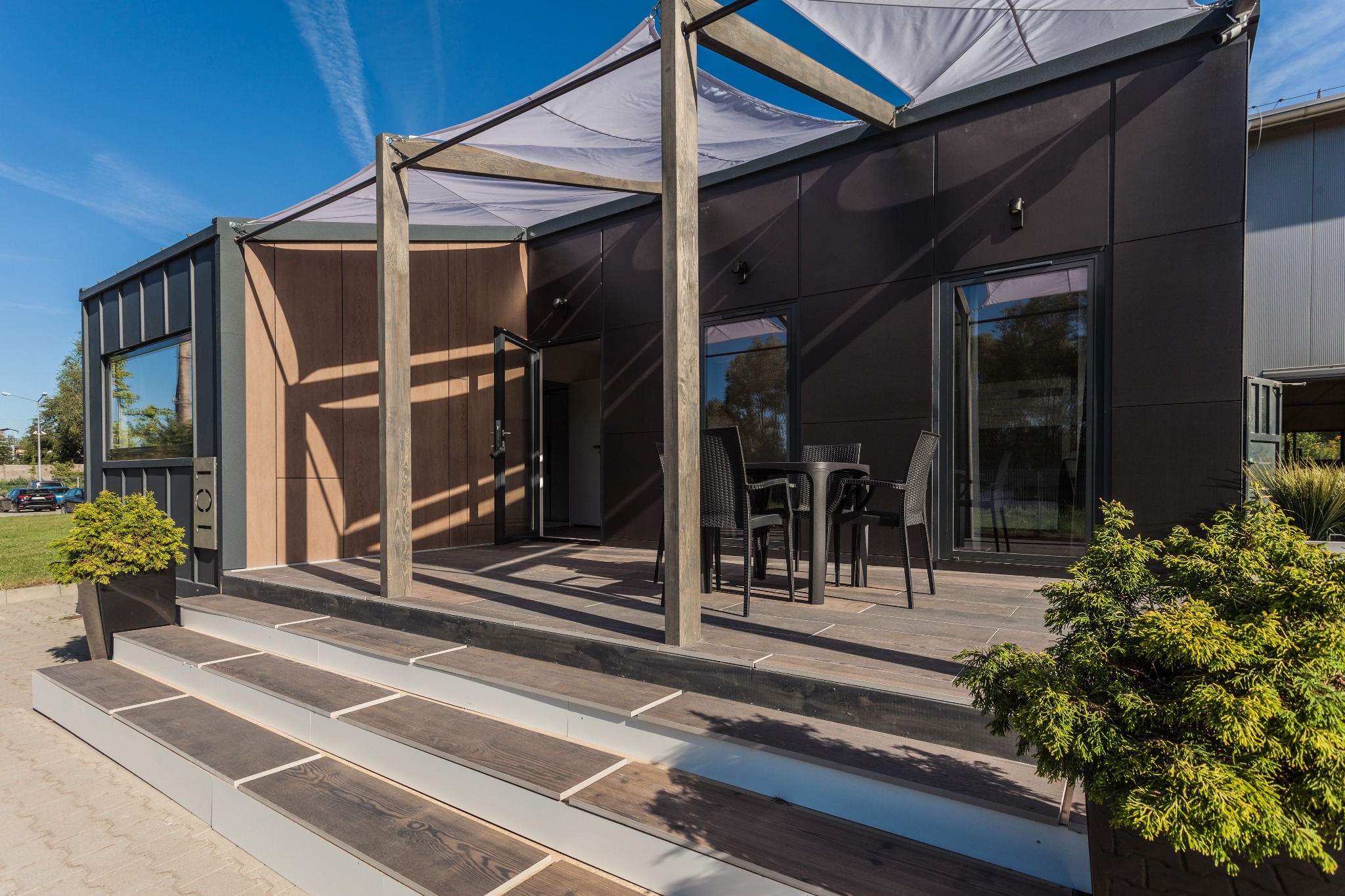 Nowoczesny dom z ciemną elewacją, drewnianym tarasem zadaszonym pergolą z szarą tkaniną, czarne meble ogrodowe i ozdobne tuje w donicach.